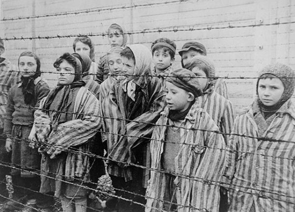 Child survivors of Auschwitz, wearing adult-size prisoner jackets, stand behind a barbed wire fence.
