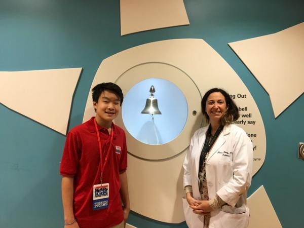 Benjamin with physician Karen Moody in front of a bell that young cancer patients ring to symbolize the end of their treatment 