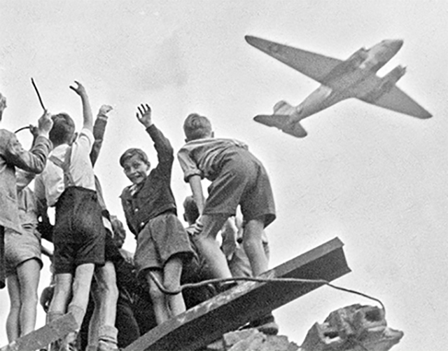 German children stand atop a ruined building in the western section of Berlin in 1948, cheering as a U.S. cargo plane arrives with food, fuel, and other supplies. Many children also got candy.